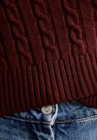 Gros plan sur un pull en maille torsadée bordeaux rentré dans un jean en denim bleu avec un bouton métallique visible.