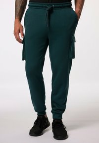 Teal Jogginghosen mit einem Kordelzugbund, seitlichen Cargo-Taschen und schmal zulaufenden Bündchen, aus weichem, glattem Stoff. Kombiniert mit schwarzen Sneakers.