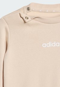 Beżowy sweter z teksturowanej tkaniny, z okrągłym dekoltem, detalami z zatrzaskami na ramieniu oraz białym logo "adidas".