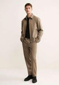 Giacca beige con zip sopra una maglietta nera, abbinata a pantaloni beige e scarpe nere. Tessuto liscio, design semplice, vestibilità dritta, accenti minimal.