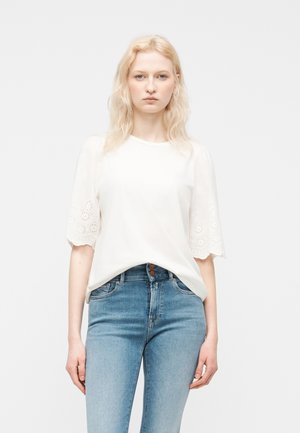 Femeie blondă cu păr ondulat, purtând o bluză albă cu mâneci decupate cu model floral și blugi din denim deschis, cu talie înaltă, pe un fundal simplu.