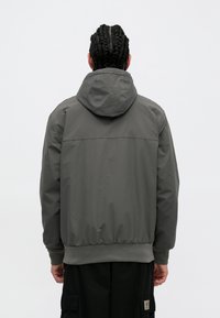 Γκρι φούτερ με κουκούλα, με ελαστικούς καρπούς και τελείωμα, προσφέροντας μια λεία υφή και έναν απλό σχεδιασμό. Η πίσω όψη αποκαλύπτει μια καθαρή σιλουέτα.