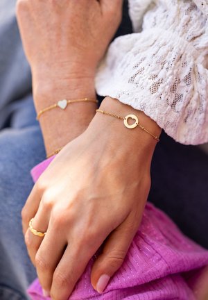 Pulseras de oro en dos muñecas que presentan un colgante de corazón y un colgante circular con un corte en forma de corazón. Las manos descansan sobre una tela texturizada de color rosa.