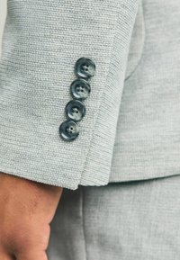 Nahaufnahme eines hellgrauen strukturierten Anzugsärmel mit vier glänzenden dunklen Knöpfen und teilweise sichtbarer Hand.