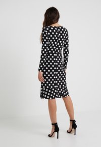 Svart klänning med vita prickar, lång ärm, figurnära design. Utsvängd nederkant med ruffle-accent. Matchas med svarta högklackade sandaler.