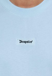 Lyseblå T-shirt lavet af blødt stof, med en ribbet rund hals og et sort mærke syet ind i forsiden med ordet "Dropsize."