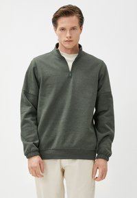 Sudadera de cuello alto con cremallera de cuarto en verde oscuro, con una superficie texturizada, puños elásticos, combinada con pantalones de color beige claro.