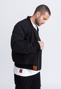 Giacca bomber nera con trama trapuntata, polsini e orlo a coste. Presenta etichette arancioni sulla parte anteriore inferiore e sulla schiena. Indossata sopra una camicia bianca.