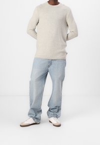Homme debout portant un pull beige texturé, un jean bleu clair à coupe décontractée et des baskets blanches avec des accents beiges sur un fond blanc uni.