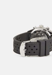 Bracelet de montre en caoutchouc noir avec un motif texturé et des perforations carrées, doté d'une boucle en métal argenté et d'une finition mate lisse.