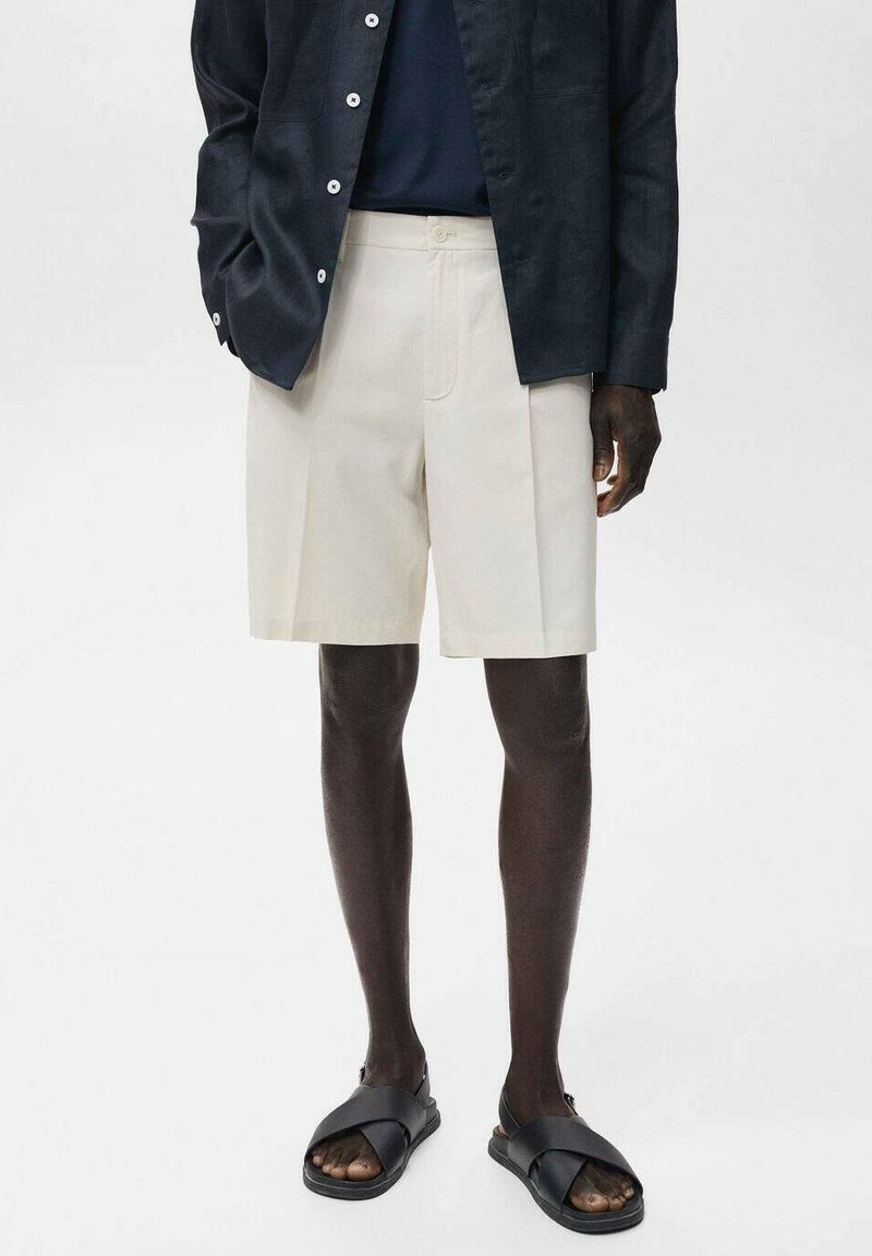 Beige skräddarsydda shorts med framfällor, tillverkade av lättviktsmaterial. Kombineras med en mörk knappad skjorta och svarta sandaler.
