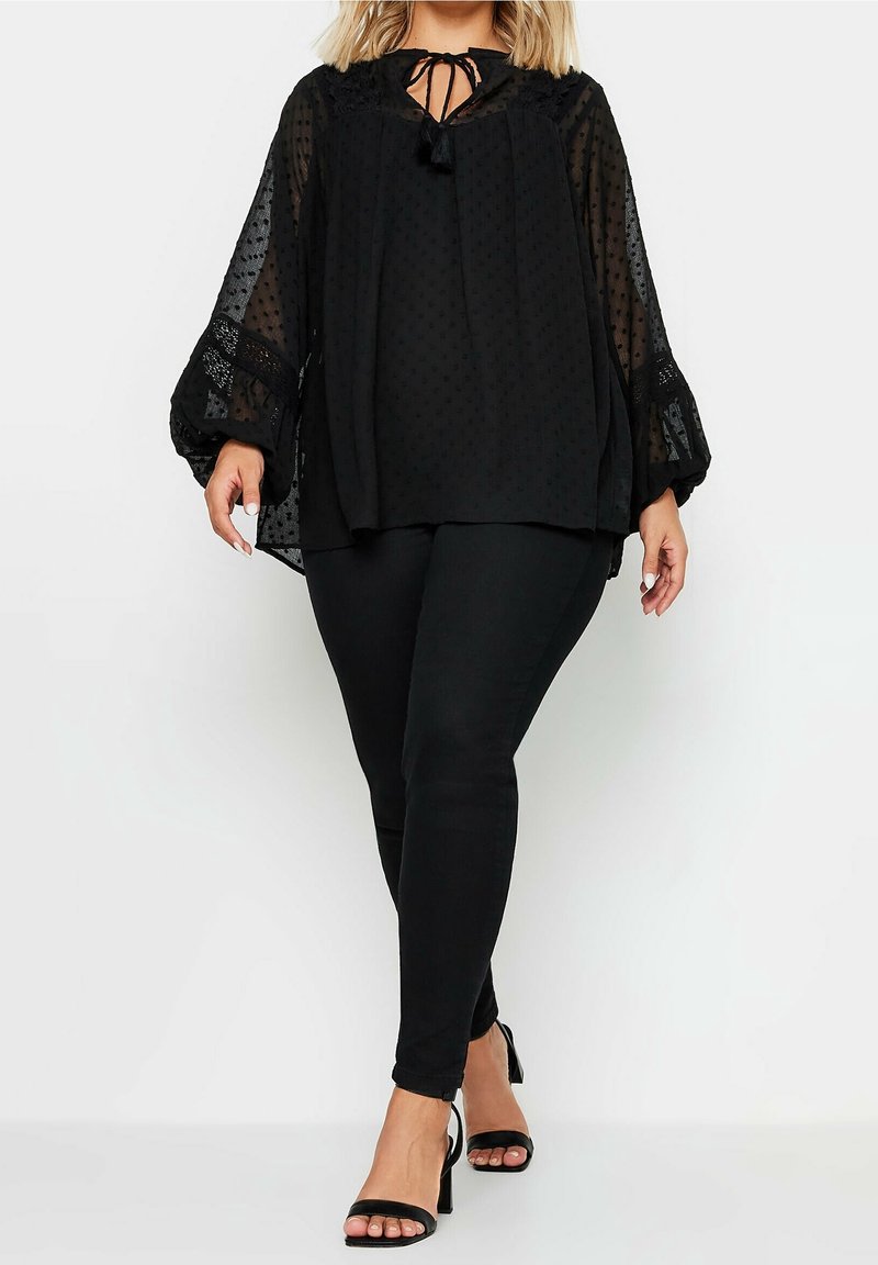 Femme portant un chemisier noir transparent à manches ballon et un pantalon noir ajusté, associé à des talons ouverts noirs, debout sur un fond blanc.