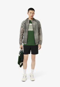 Chaqueta con capucha ligera con un patrón de rejilla negra sobre color crema; camiseta verde con logo; pantalones cortos negros; calcetines blancos; zapatillas crema; sostiene una toalla.