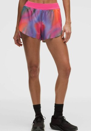 Osoba stoi i ma na sobie sportowe szorty tie-dye z różowym paskiem, czarne skarpetki i czarne trampki na jednolitym tle.