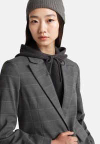 Szary marynarka w kratę z wyraźnie zarysowanymi klapami, noszona na ciemnoszarym, prążkowanym hoodie. Strój charakteryzuje się delikatną teksturą oraz detalem w postaci sznurków.