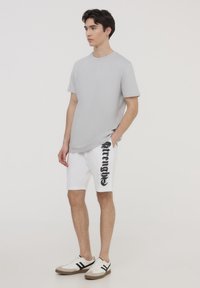 T-shirt grigia a maniche corte abbinata a pantaloni bianchi con testo grafico nero. Le scarpe da ginnastica sono bianche con strisce nere e suole color nocciola.