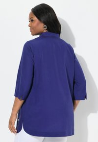 Blusa viola con colletto, maniche a tre quarti, spacchi laterali e dettaglio con bottone all'orlo. Tessuto dalla texture liscia.