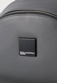 Γκρίζο σακίδιο από τεχνητό δέρμα με έντονη μαύρη τετράγωνη ετικέτα λογότυπου που γράφει "KARL LAGERFELD JEANS" σε λευκό. Διαθέτει σημαντικό φερμουάρ κλεισίματος και ραφές.