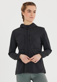 Ung kvinde med fletninger har en sort hættetrøje med lynlås og lysegrønne leggins på, stående med hænderne blidt foldet.