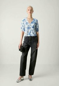 Blå blomsterkortermet bluse med v-hals, kombinert med høy midje mørke jeans. Modellen har på seg sølvfargede ballerinas og holder en liten svart håndveske.