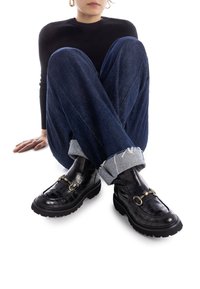 Mocasines negros con textura de cocodrilo, suela gruesa y detalle de hebilla metálica, combinados con jeans de mezclilla azul doblados y un suéter negro de canalé.