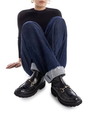 Mocasines negros con textura de cocodrilo, suela gruesa y detalle de hebilla metálica, combinados con jeans de mezclilla azul doblados y un suéter negro de canalé.