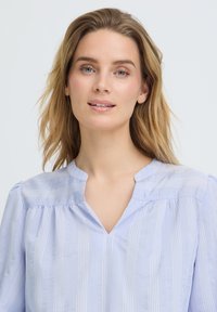 Jeune femme aux cheveux blonds portant un chemisier bleu clair à rayures avec un col en V, regardant légèrement sur le côté avec une expression neutre.
