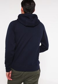 Marinblå hoodie i mjukt material, med stor huva, ribbade ärmslut och nederkant, samt en enkel, ren design utan synliga mönster.