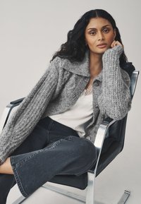 Maglione grigio lavorato a maglia con colletto, caratterizzato da tessuto texturizzato e bottoni, abbinato a una maglia bianca e jeans scuri. Modella seduta su una sedia.