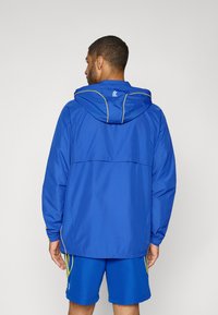 Blauwe waterdichte jas met capuchon, voorzien van gele accenten langs de naden, elastische manchetten en een ventilatieopening aan de achterkant voor ademend vermogen.