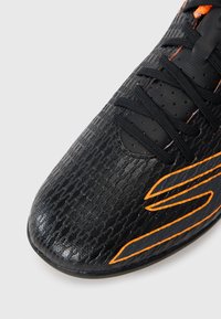 Svarta sportskor med texturerad yta, med orange accenter och svarta snörningar. Nära vy som framhäver material och design detaljer.