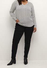 Femme portant un haut à manches longues à motifs, un pantalon noir et des bottines noires, se tenant avec une main dans la poche contre un fond uni.