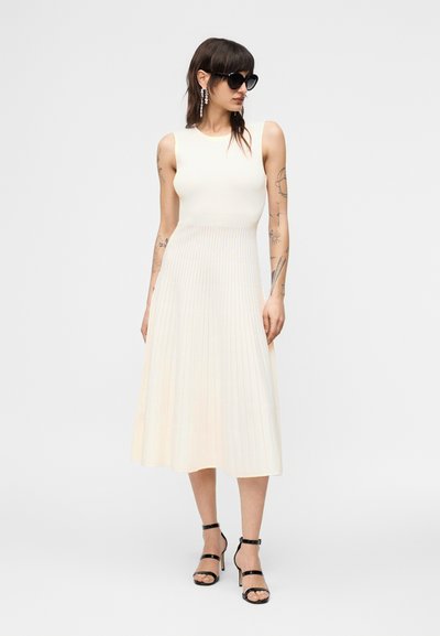 Γυναίκα με τατουάζ που φοράει λευκό πλισέ μίντι φόρεμα χωρίς μανίκια, μαύρα σανδάλια με ψηλό τακούνι, μεγάλα μαύρα γυαλιά ηλίου και κρεμαστά σκουλαρίκια.