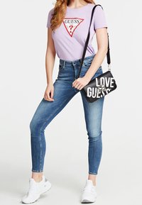 Lilla t-shirt med rødt logo, blå skinny jeans og sort håndtaske med hvid tekst. Sporty hvide sneakers fuldender outfittet.
