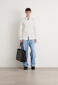 Camisa blanca de manga larga, corbata negra, jeans anchos de color azul claro y bolsa negra con "HUGO" grabado. Modelo viste zapatos negros, de pie contra una pared blanca.