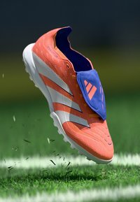 Botas de fútbol naranjas con rayas blancas, superficie texturizada y una solapa azul que cubre los cordones. Diseñadas para ofrecer tracción en el césped.