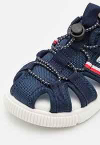 Meripihkasin tummansininen urheilumalli, jossa on avoimet varpaat, teksturoitu päällinen, joustavat nauhat ja kontrastiset raidalliset yksityiskohdat. Varusteena on valkoinen, uritettu pohja.