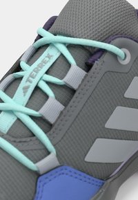 Sapatilha cinza para exterior com atacadores azul claro, detalhes em roxo e língua da marca "TERREX" em parte superior de tecido texturizado.