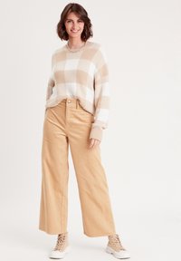 Beige und weiß kariertes Sweatshirt mit langen Ärmeln, kombiniert mit hellbraunen Cordhosen mit weitem Bein und hellen Sneakers.