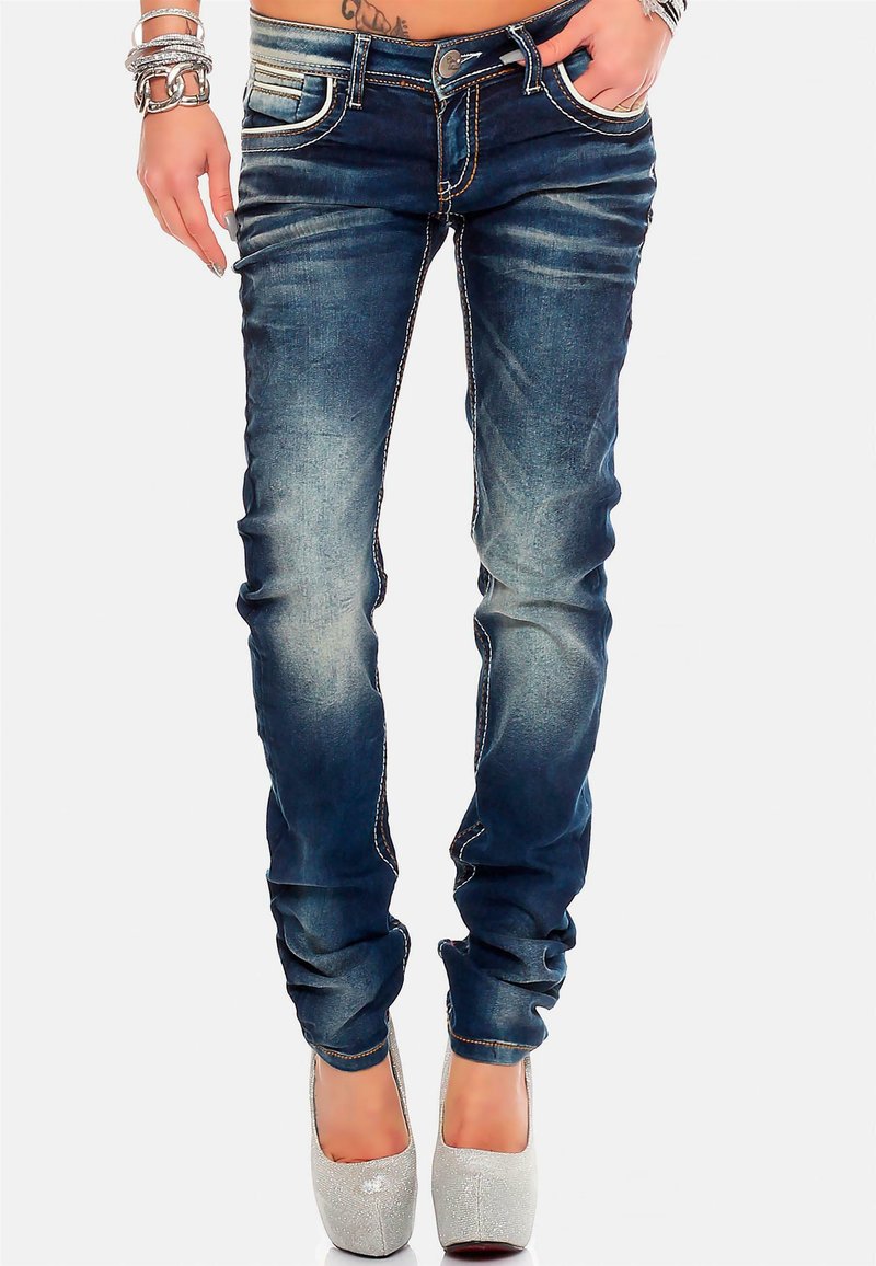 Jeans skinny in denim blu scuro con leggera scoloritura, cuciture a contrasto, cinque tasche e un bottone argentato. Indossati con tacchi alti scintillanti.