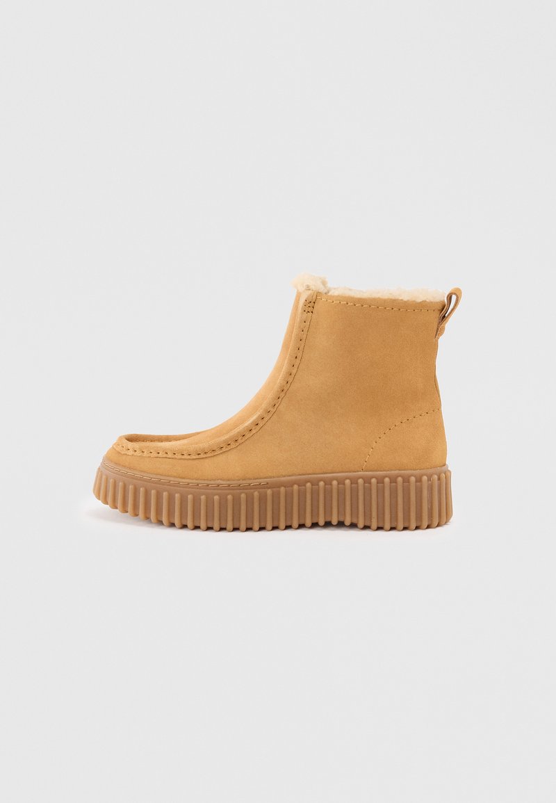 Botte de cheville en suede beige avec une doublure douce et moelleuse, un bout arrondi et une semelle en caoutchouc texturée avec des rainures verticales. Présence d'une languette à l'arrière.