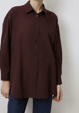 Osoba nosząca luźną, długą koszulę na guziki w ciemnobrązowym kolorze, w połączeniu z niebieskimi jeansami na jednolitym tle.