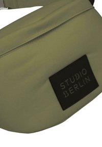 Sandy fanny pack med en jevn tekstur, med en svart lærmerkelapp med "STUDIO BERLIN" preget, og en justerbar stropp.