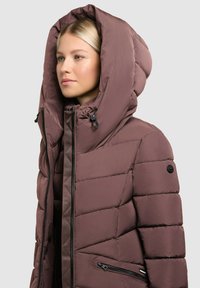 Brown puffer jacket with a large, structured hood, quilted texture, zippered front, and side pockets with black hardware accents.