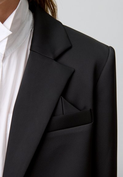 Blazer negro a medida con tejido texturizado, solapas afiladas y un bolsillo en el pecho con un pañuelo doblado de forma ordenada. Camisa blanca debajo.