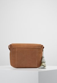 Ruskea suedeselkäinen olkalaukku, jossa on läppäsuljin ja harmaasta ja vihreästä kolmekerroksisesta tupsusta koostuva koriste sivussa, valkoisella jalustalla.