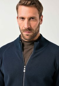 Námořnický svetr na zip s vysokým límcem, žebrovanými detaily a hladkou texturou, nošený přes hnědou košili. Zip má stříbrný táhlo.