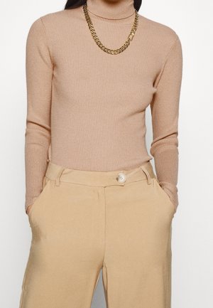 Personne portant un pull à col roulé côtelé beige, un pantalon beige avec une fermeture par bouton et un collier chaîne dorée épais.
