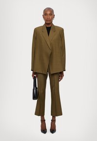 Costum pe verde măsliniu, croi ajustat, cu blazer structurat și pantaloni evazați, având un finisaj texturat. Accesorizat cu o geantă de mână neagră și pantofi cu toc.