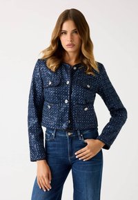 Donna con capelli castano chiaro ondulati indossa una giacca di paillettes blu navy con bottoni argentati e jeans blu scuro, in posa con una mano sulla vita.